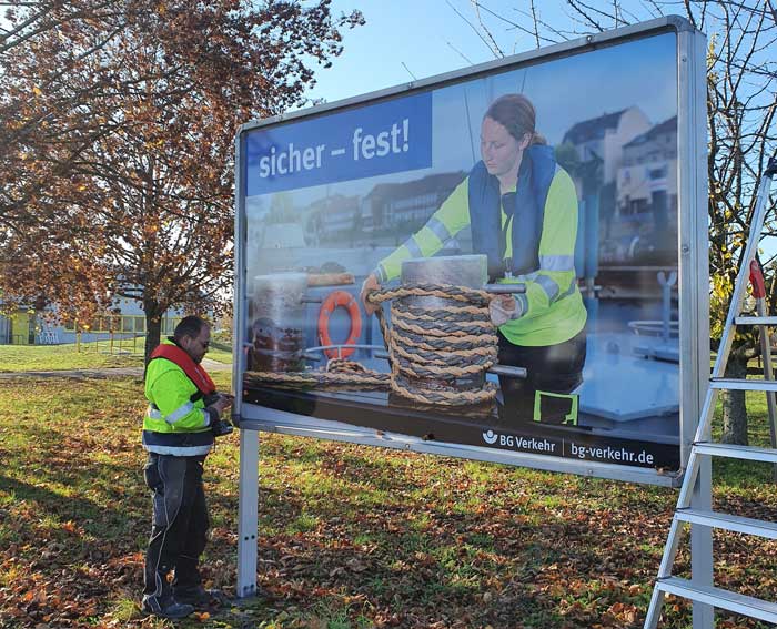 Arbeiter steht neben neuem Plakat an BG-Werbetafel